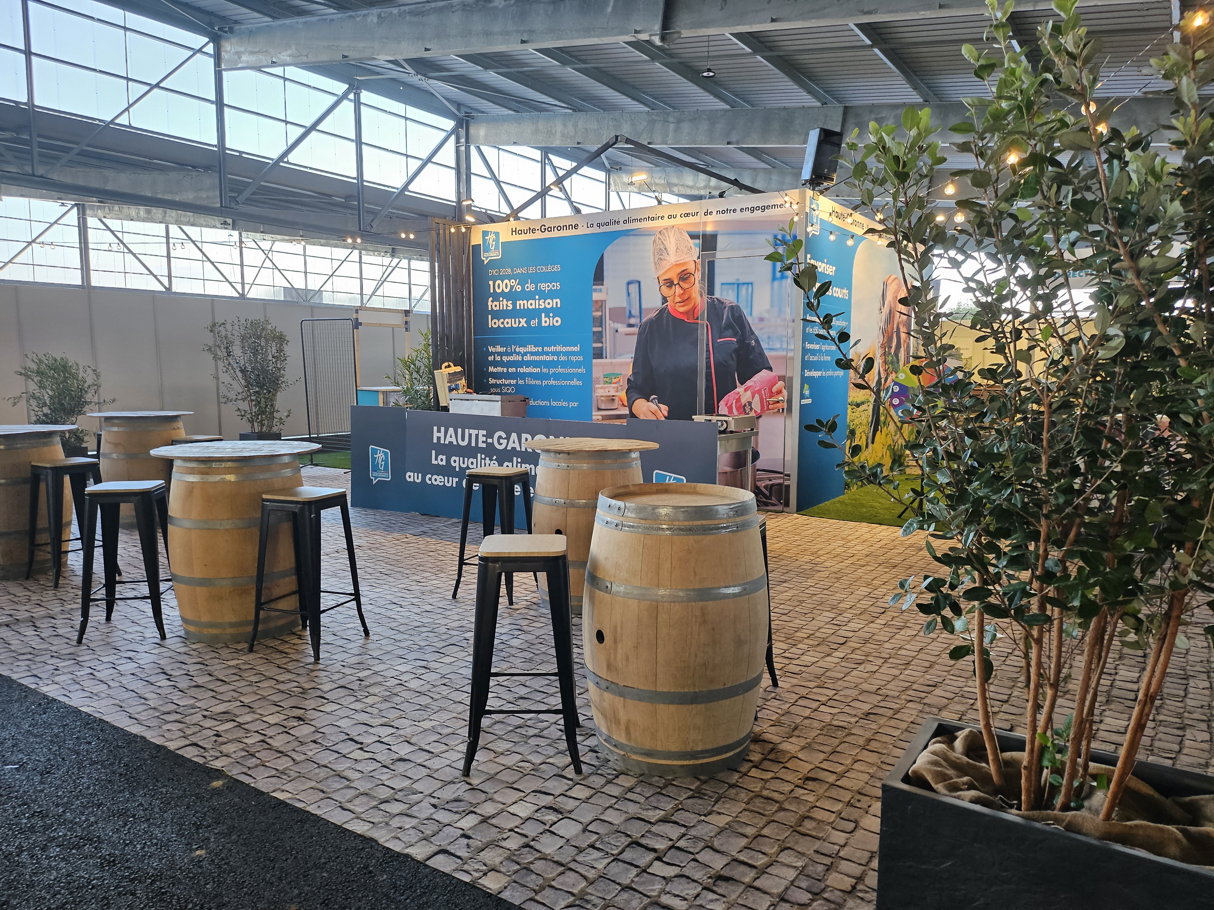 stand du département de la Haute-Garonne au salon Les Pyrénéennes, avec des tonneaux en guise de tables hautes et tabouret sur un sol imitation pavé